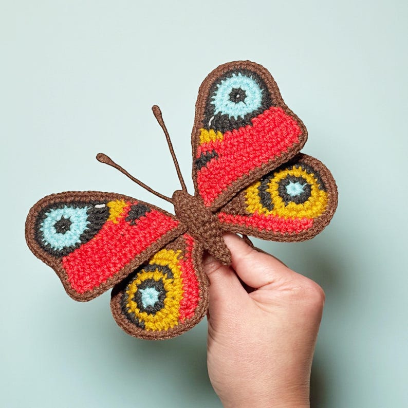 May include: A crocheted butterfly ornament featuring a brown body and antennae. The wings are detailed with vibrant red, yellow, and light blue circular patterns, all outlined in a dark brown yarn border, creating a textured and colorful appearance.