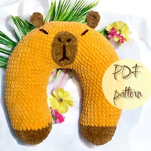 May include: A U-shaped plush pillow designed to resemble a capybara, featuring a golden-yellow textured body, dark brown snout, ears, and feet, with closed eyes. A circular overlay on the right side displays the text PDF pattern in brown script.