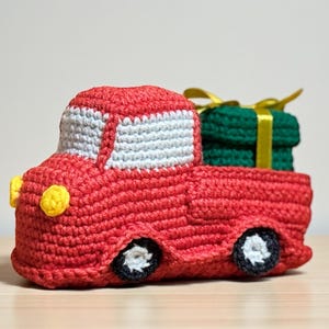 Könnte beinhalten: Ein handgefertigter roter Häkellastwagen mit weißer Kabine und gelben Scheinwerfern. Die Ladefläche des Lastwagens enthält ein grünes Geschenk, das mit einem gelben Band verpackt ist. Die Räder sind schwarz mit weißen Zentren. Eine charmante, festliche Dekoration.