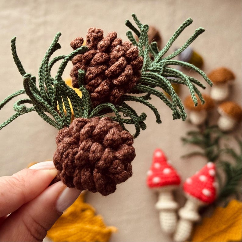 Pode incluir: Close-up de pinhas marrons de croch&ecirc; com detalhes verdes. No fundo, cogumelos de croch&ecirc; com chap&eacute;us vermelhos e brancos e outros itens de croch&ecirc; com tema de outono.