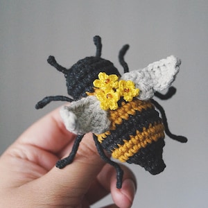 May include: A crocheted bee figurine featuring a black head and antennae, a striped black and yellow abdomen, and light grey wings. The bee is decorated with three small yellow crocheted flowers with brown centres attached to its back.