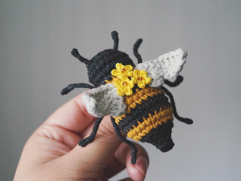 May include: A crocheted bee figurine featuring a black head and antennae, a striped black and yellow abdomen, and light grey wings. The bee is decorated with three small yellow crocheted flowers with brown centres attached to its back.