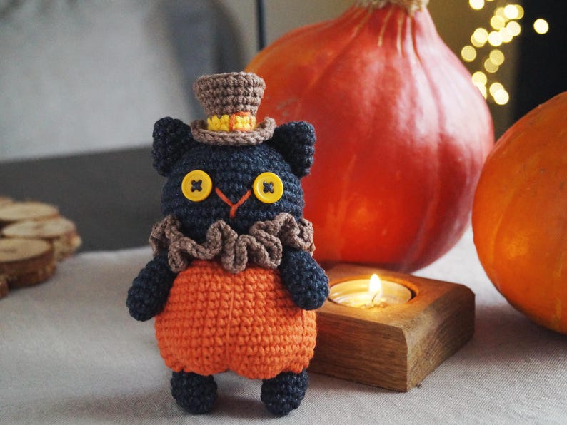 May include: A handmade crochet cat doll with a top hat, ruffled collar, and orange pumpkin-shaped body. The cat has button eyes and is set against a backdrop of pumpkins and a lit candle, evoking a Halloween theme.