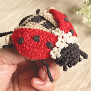 May include: A crocheted ladybird figurine featuring a bright red shell with black spots, a dark grey head with black bead eyes, and small white crocheted flowers with gold centres attached to the side. The insect has thin black crocheted legs.