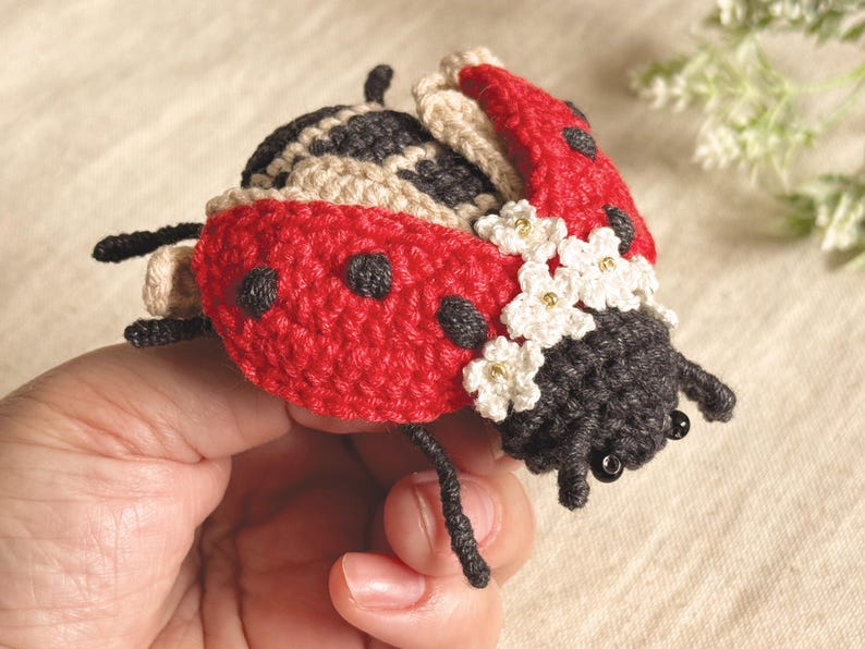 May include: A crocheted ladybird figurine featuring a bright red shell with black spots, a dark grey head with black bead eyes, and small white crocheted flowers with gold centres attached to the side. The insect has thin black crocheted legs.