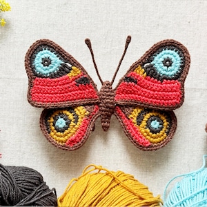May include: A crocheted butterfly applique featuring a brown body and antennae. The wings display intricate patterns in vibrant red, bright yellow, light blue, and dark brown yarn, with circular eye-like motifs on each of the four wing sections.
