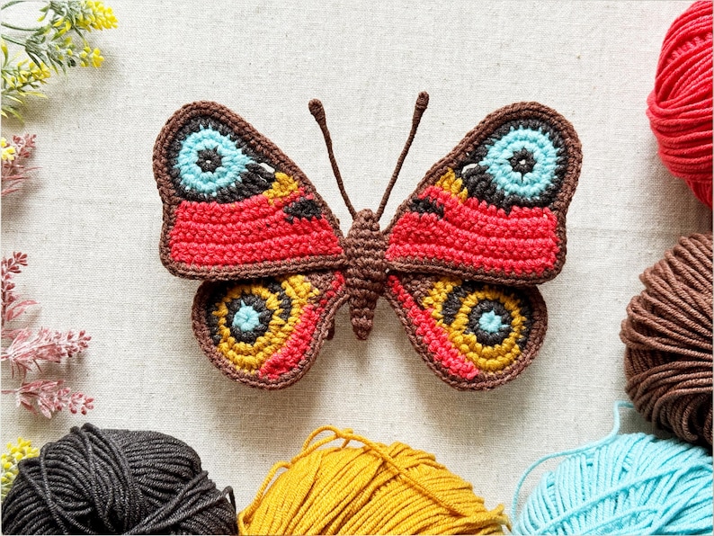 May include: A crocheted butterfly applique featuring a brown body and antennae. The wings display intricate patterns in vibrant red, bright yellow, light blue, and dark brown yarn, with circular eye-like motifs on each of the four wing sections.