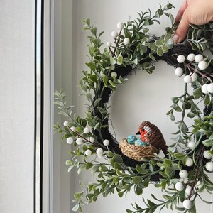 May include: A decorative wreath featuring a crocheted bird sitting on a nest with blue eggs. The wreath is adorned with green leaves and white berries, creating a natural and inviting aesthetic. The wreath is held by a hand.