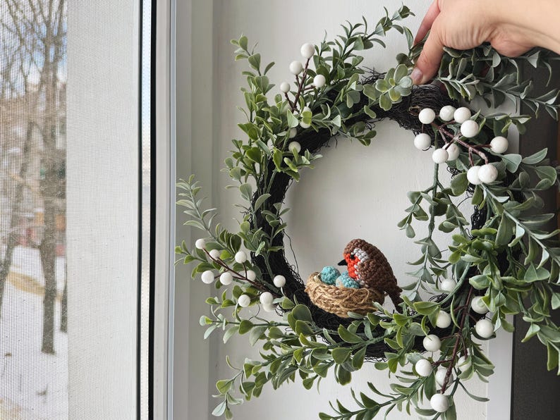May include: A decorative wreath featuring a crocheted bird sitting on a nest with blue eggs. The wreath is adorned with green leaves and white berries, creating a natural and inviting aesthetic. The wreath is held by a hand.