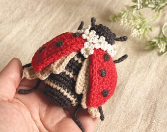 May include: A handmade crochet ladybug with a red shell, black spots, and a black head. The ladybug has a white flower crown and is held in a hand. The ladybug is approximately 3 inches long.