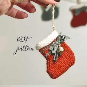 Könnte beinhalten: Ein kleiner, gestrickter Weihnachtsstrumpf-Ornament in Rot-, Weiß- und Grautönen, mit einer kleinen goldenen Glocke und einem grauen Band. Der Text "PDF pattern" ist in schwarzer Schrift geschrieben. Das Ornament wird von einer beigen Schnur gehalten.