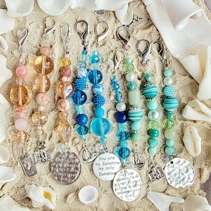 May include: Six colorful beaded keychains with charms featuring nautical themes and inspirational messages. The charms include a seashell, a sailboat, a lighthouse, and a heart. The messages on the charms include "May you always have a shell in your pocket & sand between your toes", "You are my only Sunshine", and "May you always have a shell in your pocket & sand between your toes".