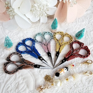 May include: A collection of decorative embroidery scissors in various colors, including blue, pink, gold, and red. The scissors have ornate handles and are arranged in a fan shape. A charm with a snowman, snowflake, and reindeer is also present.