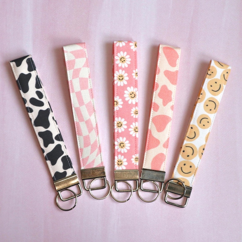 May include: Five colorful keychains with different patterns, including cow print, checkered, daisies, abstract shapes, and smiley faces. Each keychain has a silver key ring.