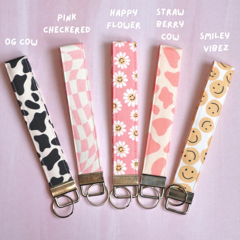 May include: Five different keychains with various patterns. The keychains are pink, white, and black with designs of cows, flowers, smiley faces, and checkered patterns. The keychains have the text "OG Cow", "Pink Checkered", "Happy Flower", "Straw Berry Cow", and "Smiley Vibez" written above them.