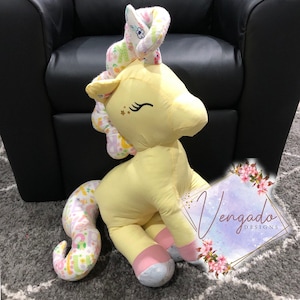 May include: A yellow unicorn plush toy with a pink and white floral patterned tail and horn. The unicorn has a pink nose and pink feet. The unicorn is sitting on a gray carpet. The image also includes a pink and white floral design with the text "Vengado Designs".