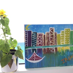 Puede incluir: Una pintura colorida de un horizonte urbano con un barco en el agua. La pintura está sobre un lienzo y tiene un fondo azul con estrellas blancas. Los edificios de la ciudad están pintados en varios colores, incluyendo morado, verde, amarillo y marrón. El barco está pintado en rojo y blanco. La pintura está firmada "TREYW" en la esquina inferior derecha.