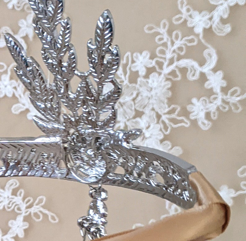 May include: A silver headband with a decorative floral design and a dangling crystal charm. The headband is on a beige background.