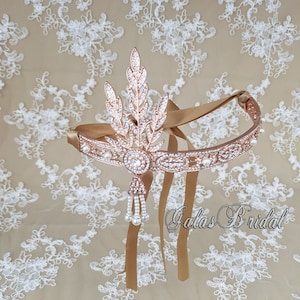May include: Rose gold bridal headband with a pearl and crystal design. The headband has a leaf motif and a long pearl and crystal tassel. The headband is on a white lace background. The text "Galas Bridal" is visible in the bottom right corner of the image.