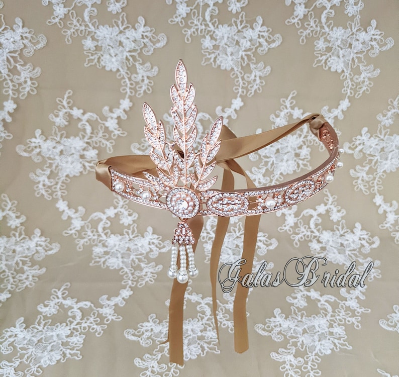 May include: Rose gold bridal headband with a pearl and crystal design. The headband has a leaf motif and a long pearl and crystal tassel. The headband is on a white lace background. The text "Galas Bridal" is visible in the bottom right corner of the image.