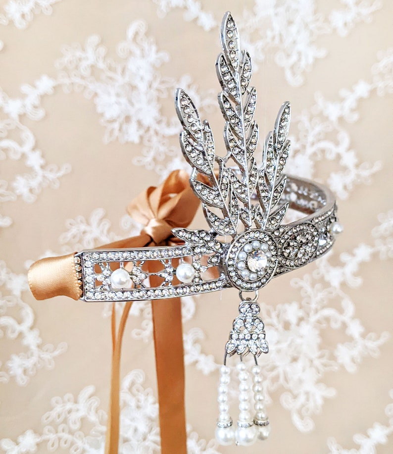 May include: A silver headband with a delicate floral design and pearl accents. The headband is adorned with rhinestones and has a dangling pearl and rhinestone drop.