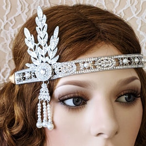 May include: A silver headband with a pearl and rhinestone design. The headband has a leaf-shaped design on one side and a dangling pearl chain on the other.