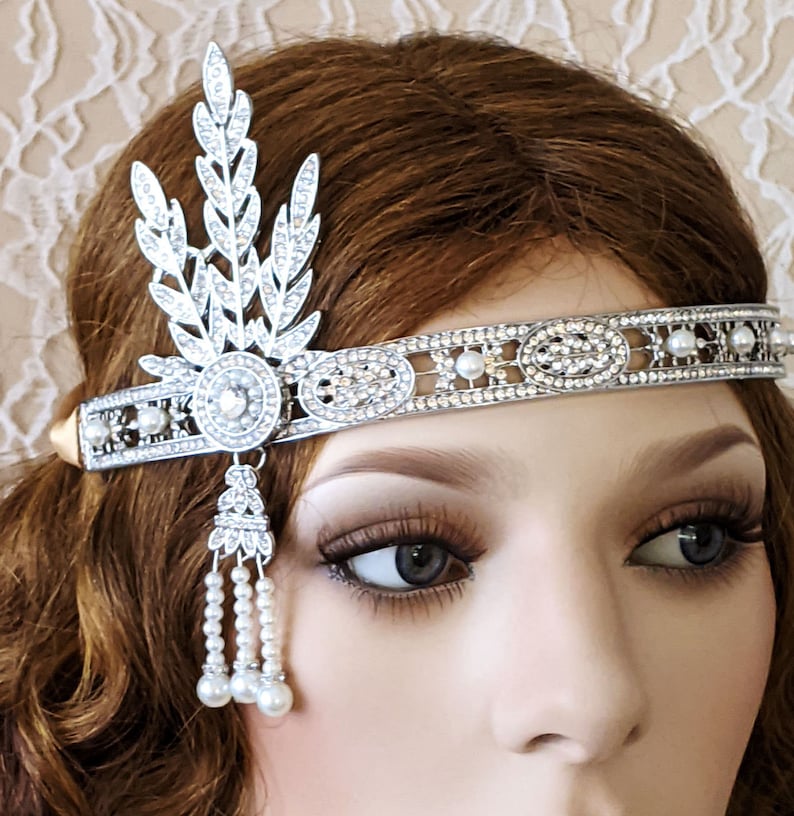 May include: A silver headband with a pearl and rhinestone design. The headband has a leaf-shaped design on one side and a dangling pearl chain on the other.