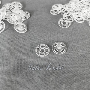 May include: A collection of clear plastic snap fasteners scattered on a gray fabric surface. The snaps are circular with multiple holes and a central stud. The text "Gala's Bridal" is embroidered on the fabric.
