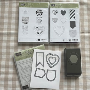 May include: Two clear plastic packaging sheets with black and white stamp designs. The designs include hearts, banners, and text such as "Merry Christmas to You" and "Happy Valentines Day". A white sheet with black and white cut-out designs of hearts and banners. A black and gray punch tool with a hexagonal hole.