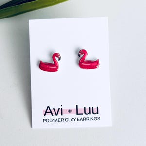 May include: A pair of bright pink flamingo pool float earrings attached to a white display card. The card features the black text Avi + Luu and the words POLYMER CLAY EARRINGS printed below. The flamingo earrings are shaped like inflatable rings.