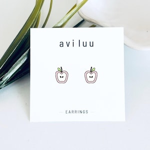 May include: A pair of stud earrings featuring a small, light pink apple slice design with a green leaf and black seeds. The earrings are displayed on a white square card with the text aviluu and EARRINGS printed in black sans-serif font.