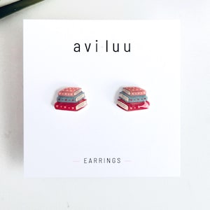 Peut inclure: Une paire de boucles d'oreilles sur un support blanc marqué 'avi luu' et 'EARRINGS'. Chaque boucle représente une pile de trois livres dans des tons rose, bleu et rouge, ornés de petits motifs de cœurs sur les couvertures et les tranches.