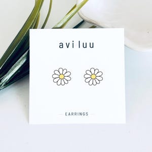 Op de afbeelding: Een paar oorknopjes met een wit madeliefje-ontwerp met een geel hartje, gepresenteerd op een witte vierkante kaart met de tekst aviluu en EARRINGS in het zwart gedrukt. De oorbellen liggen op een witte ondergrond.