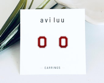 Puede incluir: Un par de pendientes de botón con una letra O mayúscula roja con borde blanco, montados sobre una tarjeta de presentación blanca con el nombre de la marca aviluu y la palabra EARRINGS en texto negro. El diseño es sencillo y geométrico.