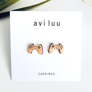 May include: A pair of stud earrings shaped like tan video game controllers with black buttons and red accents, displayed on a white card with the text aviluu and EARRINGS printed in black. The earrings feature a glossy finish and detailed graphics.