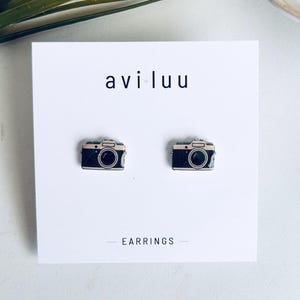 May include: A pair of stud earrings shaped like vintage cameras with black and silver details. The earrings are attached to a white display card featuring the text avi luu and EARRINGS printed in black ink on the front of the card.
