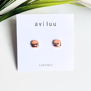 May include: A pair of stud earrings featuring a miniature hamburger design with a tan bun, green lettuce, and dark brown patty. The earrings are displayed on a white card with the text "avi luu" and "EARRINGS" printed in black.