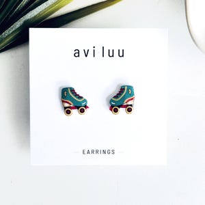 May include: A pair of stud earrings shaped like roller skates on a white display card with the text aviluu and EARRINGS. The skates are teal with red laces, yellow lightning bolt accents, and red wheels with black centres, featuring a flat design.