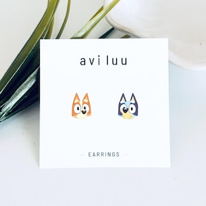 May include: A pair of stud earrings featuring the cartoon dog characters Bingo and Bluey on a white display card. The card displays the text aviluu at the top and EARRINGS at the bottom. The characters are depicted with their signature orange and blue colours.