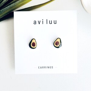May include: A pair of stud earrings shaped like avocado halves, featuring dark green skin, light yellow flesh, and a dark red pit. The earrings are mounted on a white display card with the text avi luu and EARRINGS printed in black.