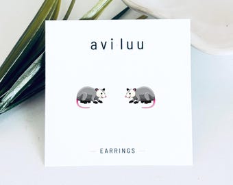 May include: A pair of stud earrings featuring a cartoon opossum design on a white display card. The card displays the text avi luu at the top and EARRINGS at the bottom. Each opossum is grey with a white face, black feet, and a pink tail.