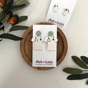 May include: White polymer clay earrings in the shape of a medical scrubs top with a stethoscope design. The earrings have a light green circular top and are displayed on a white card with the text "Avi + Luu Polymer Clay Earrings". A matching pair of stud earrings is also shown.