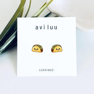 May include: A pair of stud earrings featuring a cartoon taco design with a smiling face, pink cheeks, and yellow shell, displayed on a white square card with the text aviluu and EARRINGS printed in black, centered on the card surface.