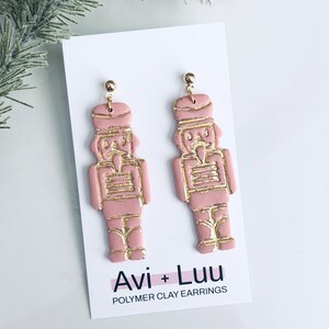 May include: A pair of pink polymer clay earrings shaped like nutcrackers with gold detailing. The earrings are presented on a white card, which features the text "Avi + Luu" and "Polymer Clay Earrings".