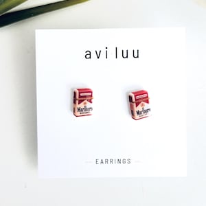 Peut inclure: Une paire de boucles d'oreilles en forme de paquets de cigarettes miniatures rouges et blancs avec le texte Marlboro Red Label imprimé sur le devant. Les boucles d'oreilles sont fixées sur une carte de présentation blanche avec le texte avi luu et EARRINGS.