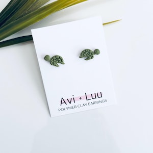 May include: Olive green polymer clay turtle earrings on a white card with the text "Avi + Luu POLYMER CLAY EARRINGS". The earrings are small, detailed, and turtle-shaped. Green plant stems are in the background.