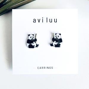 May include: A pair of stud earrings featuring a black and white panda bear illustration holding a piece of bamboo. The earrings are mounted on a white display card with the text avi luu and EARRINGS printed in black ink on the front surface.