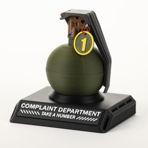 Può includere: Un accessorio da scrivania originale a forma di granata verde. Presenta una base nera con la scritta "COMPLAINT DEPARTMENT TAKE A NUMBER" e un'etichetta gialla con il numero "1".