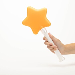 Peut inclure: Une baguette magique en forme d'&eacute;toile avec une t&ecirc;te orange et un manche blanc. La baguette est tenue dans une main sur fond blanc. L'&eacute;toile est d'un orange vif et uni.