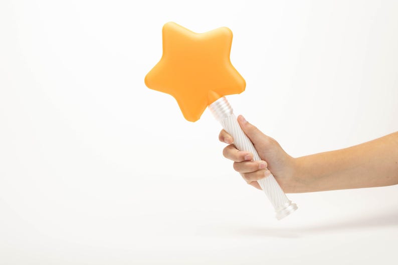 Peut inclure: Une baguette magique en forme d'&eacute;toile avec une t&ecirc;te orange et un manche blanc. La baguette est tenue dans une main sur fond blanc. L'&eacute;toile est d'un orange vif et uni.